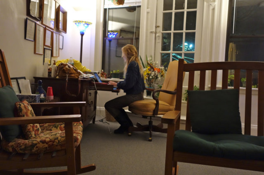A woman works on a laptop at a desk in a cozy office, surrounded by plants and framed documents.