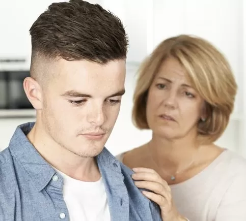 A woman comforts a young man, who appears distressed, in a bright kitchen setting.