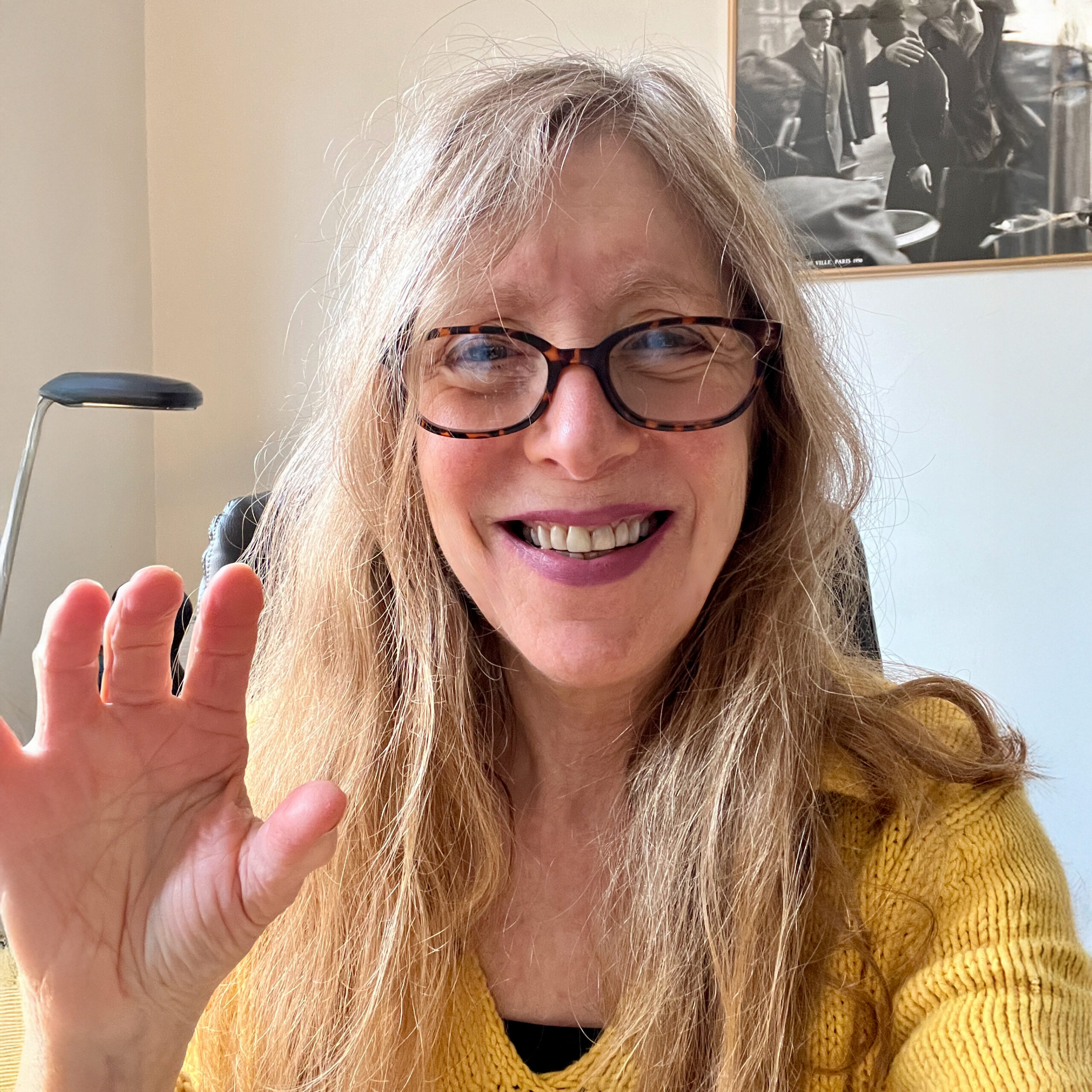 Dr. Lynn Margolies smiles and waves, wearing a yellow sweater and glasses, in a well-lit room.
