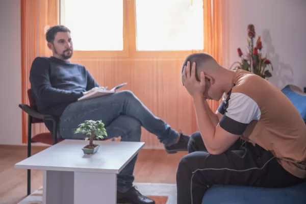 A male therapist listens as a teenage boy sits with his head in his hands, expressing distress in a cozy room.