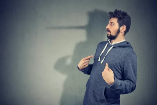 A man in a hoodie points to his shadow, which resembles a long nose, against a gray wall.
