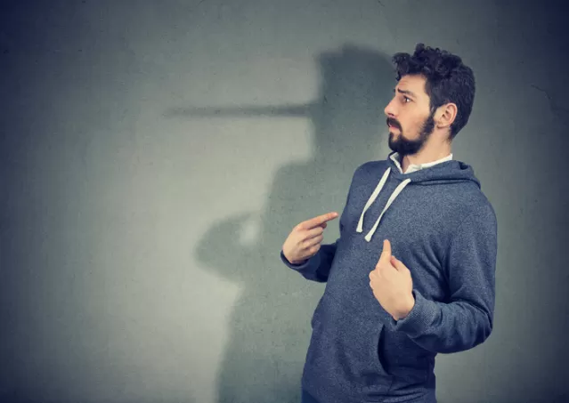 A man in a hoodie points to his shadow, which resembles a long nose, against a gray wall.