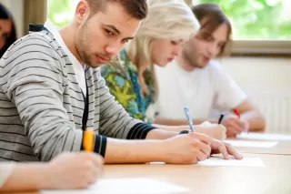 Four students sit at a table, focused on writing during a test.