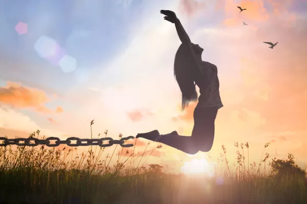 A woman jumps joyfully against a sunset, breaking free from chains, with birds flying in the sky.