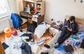 A person sits on a bed, using a device, surrounded by scattered clothes and a messy room.