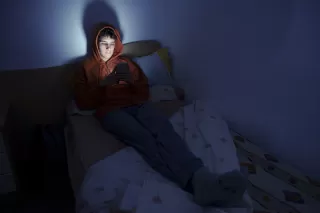 A person in a hoodie sits on a bed, looking at a phone in a dimly lit room.