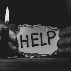 A person holds a torn paper with "HELP" written on it, illuminated by a small flame.