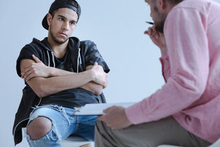 A young man sits with crossed arms, listening intently to a man in a pink shirt, who holds a clipboard.