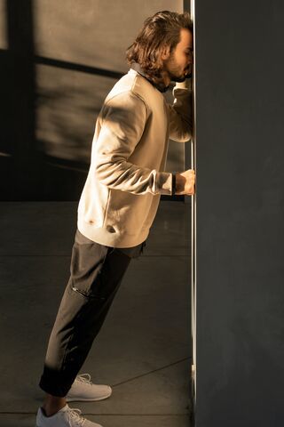 A man leans against a wall, resting his forehead on it, wearing a light sweater and dark pants.