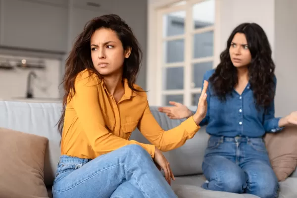 Two women sit on a couch, one looks upset and turns away, while the other gestures expressively, appearing frustrated.